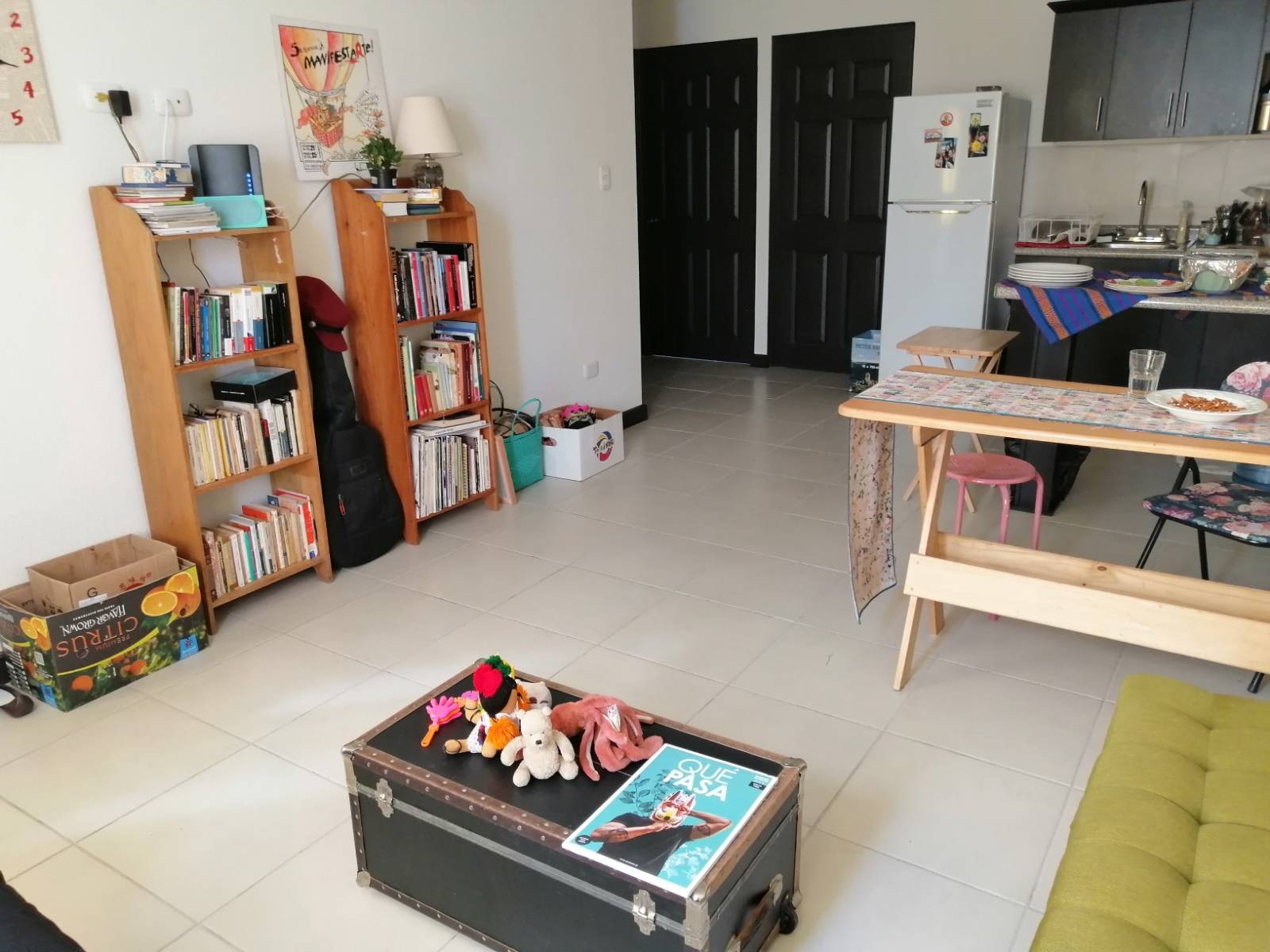 A Living Room with a table, bookshelves, magazines on a table top, and a kitchen in the background